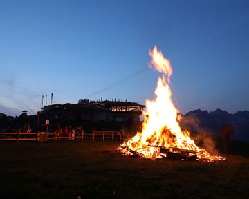 Ein großes Feuer brennt auf einer Wiese bei Dämmerung. Im Hintergrund sind Menschen und eine Gebäudeansicht sichtbar.