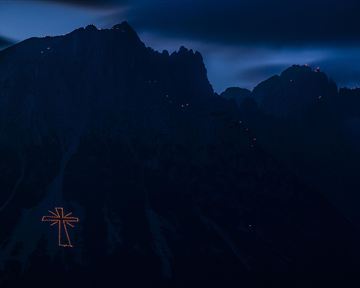 Ein großes, beleuchtetes Kreuz auf einem Berg im Dunkeln. Die Umgebung ist rau und gebirgig, mit dunklen Wolken am Himmel.