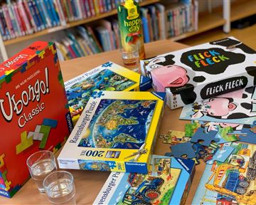 A table with board games and puzzles is located in a library. Various game boxes and puzzle pieces are visible.