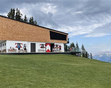A modern building with a sloped roof structure stands on a meadow. In the background, there are mountains and a cloudy sky.