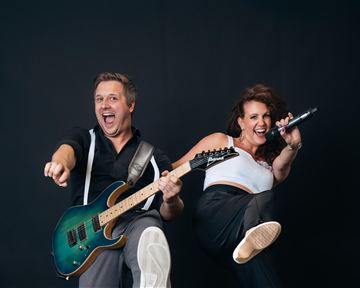 A musician with a guitar and a singer are happily posing in front of a neutral background. Both radiate energy and joy.