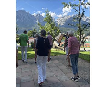 A group of older people is walking in a beautiful landscape with mountains in the background. The sky is blue and the surroundings are green.