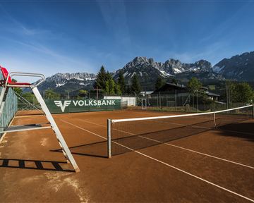 Ein Tennisplatz mit rotem Sand und einer imposanten Bergkulisse im Hintergrund. Die Sonne scheint und der Himmel ist klar.