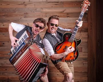 Two musicians happily pose in front of a wooden wall. One plays the accordion and the other holds a guitar.