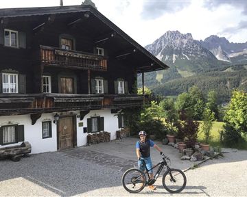 Ein Radfahrer steht vor einem traditionellen Chalet in den Bergen. Im Hintergrund sind grüne Wiesen und beeindruckende Berggipfel zu sehen.