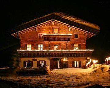 Ein schönes Holzhaus im Schnee, das festlich mit Lichtern dekoriert ist. Fackeln beleuchten den Weg zum Eingang und schaffen eine warme Atmosphäre in der Nacht.