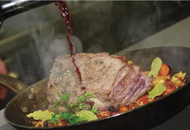 Ein saftiges Stück Fleisch wird in einer Pfanne mit Gemüse zubereitet. Aromatischer Dunst steigt auf, während Rotwein hinzugefügt wird.