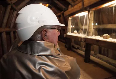 Eine Person mit einem weißen Helm und einem silbernen Mantel steht in einem Raum mit Vitrinen. In den Vitrinen sind verschiedene Objekte zu sehen.