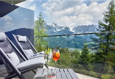 Eine gemütliche Terrasse mit zwei Liegestühlen und einer atemberaubenden Aussicht auf die Berge. Auf dem Tisch stehen zwei bunte Cocktails.