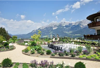 Eine schöne Gartenlandschaft mit bunten Pflanzen und einem Wasserbecken. Im Hintergrund sieht man beeindruckende Berge unter einem blauen Himmel.