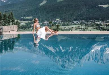 A woman relaxes at the edge of a swimming pool with a view of majestic mountains. The water reflects the surroundings and the sky is clear and blue.