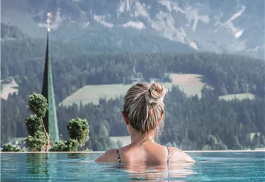 A woman stands in the water of a pool and looks at the mountains. The landscape is picturesque with green meadows and a church in the background.