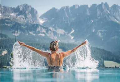 A woman in the water of an infinity pool enjoys the view of the mountains. Splashes of water surround her and reflect the beauty of nature.