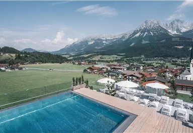 A breathtaking view of the mountains and a swimming pool on a terrace. In the background, picturesque houses and a church can be seen.