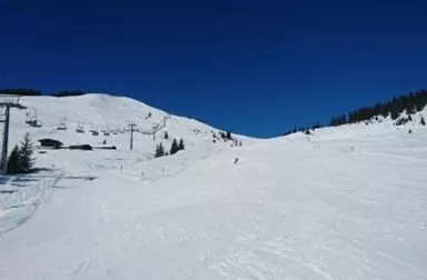 Eine schneebedeckte Skipiste unter einem klaren blauen Himmel. Im Hintergrund sind Bäume und ein Skilift zu sehen.