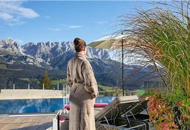 Eine Person in einem Bademantel steht am Rand eines Pools und blickt auf bergige Landschaft. Der Himmel ist klar und die Umgebung wirkt entspannt und einladend.