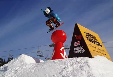 Ein Snowboarder springt über eine rote Kuppel auf einer schneebedeckten Piste. Im Hintergrund sind Skilifte und eine Werbetafel sichtbar.