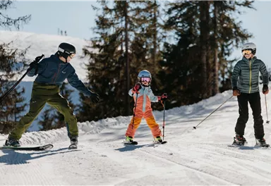 Eine Familie fährt Ski in einer sonnigen Winterlandschaft. Das Kind lernt das Skifahren, während die Erwachsenen zuschauen und helfen.