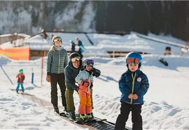 Eine Gruppe von Kindern steht in einem verschneiten Skigebiet, während sie auf den Skilift warten. Die Sonne scheint und die Atmosphäre ist fröhlich und spielerisch.