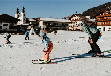 Ein Kind und ein Erwachsener skifahren in einer verschneiten Landschaft. Im Hintergrund sind traditionelle Gebäude und eine Kirche zu sehen.