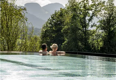 Ein Paar entspannt in einem Infinity-Pool mit Blick auf die Berge. Die Umgebung ist grün und die Sonne scheint.