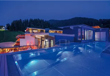 A modern building with a lit pool at night. In the background, trees and hills can be seen.