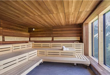 A modern sauna interior with wooden paneling and large windows. The view overlooks an impressive mountain landscape.