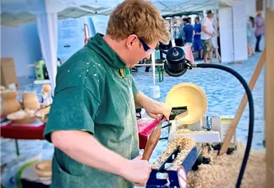 A craftsman works on wood at a lathe. In the background, stalls and people can be seen at a market.