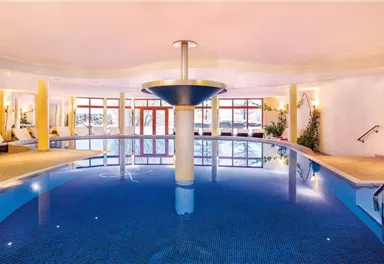 Ein eleganter Innenpool mit blauem Wasser und einer modernen Wasserfontäne. Die Umgebung ist hell und einladend, mit Fenstern, die natürliches Licht hereinlassen.