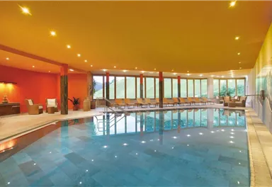 A modern indoor swimming pool with smooth water and comfortable loungers. Large windows provide a view of the green surroundings.