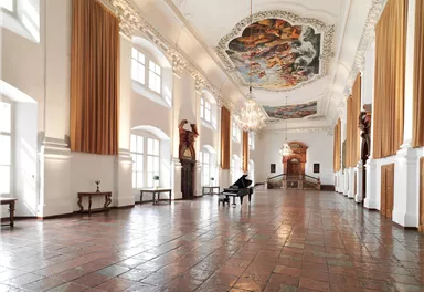 An elegant room with high ceilings and an impressive fresco. At the center is a piano, surrounded by windows and decorative elements.