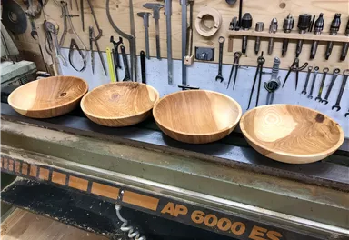 Four wooden bowls in different colors are on a work table. In the background, numerous tools are hanging on the wall.