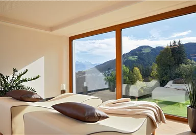 Ein gemütlicher Raum mit zwei Liegen und großen Fenstern. Im Hintergrund sieht man eine malerische Berglandschaft unter einem klaren Himmel.