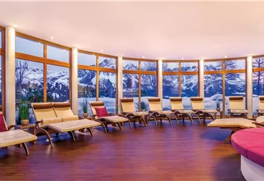 Ein helles Entspannungszimmer mit bequemen Liegen und großen Fenstern. Im Hintergrund sind schneebedeckte Berge und ein schöner Sonnenuntergang zu sehen.