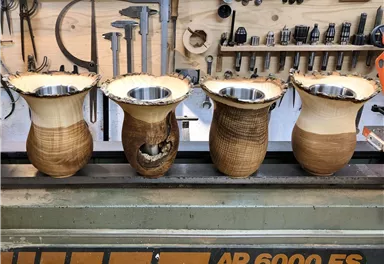 Four intricately crafted wooden vases stand on a workbench. In the background, various tools and materials are visible.