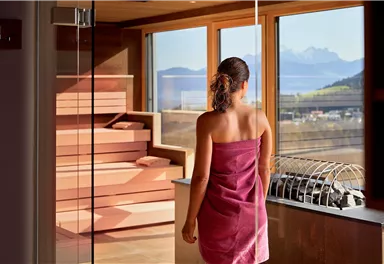 Eine Frau in einem Handtuch schaut aus einem großen Fenster auf die Berge. Der Raum ist modern und ruhig, mit Holz- und Glaselementen.