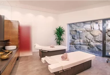 A modern wellness room with two massage tables and a large window overlooking a stone wall. The decor is minimalist, featuring plants and elegant wooden elements.