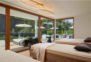 Ein modernes Schlafzimmer mit zwei Betten und großen Fenstern. Draußen sind Liegen und Sonnenschirme sichtbar, umgeben von Natur.