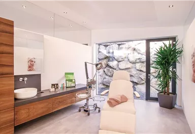 A modern treatment room with a couch and a large window that overlooks a stone wall view. The room is bright and minimalist in design, with plants and some cosmetic products.