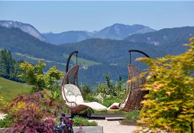 Ein schöner Garten mit zwei hängenden Stühlen und einer malerischen Berglandschaft im Hintergrund. Die Pflanzen umgeben die Sitzecke und schaffen eine entspannende Atmosphäre.