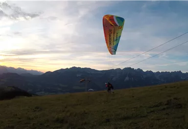 Ein Paraglider schwebt über einer Berglandschaft bei Sonnenuntergang. Der Himmel ist bunt und die Berge sind im Hintergrund sichtbar.