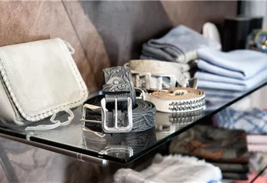 An elegant selection of accessories on a glass shelf. On display are a handbag, several belts, and folded scarves.