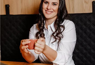 A woman with long hair is sitting at a table holding a cup. In front of her is a small dessert on a plate and a laptop is on the table.