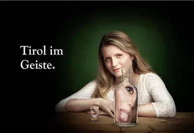 A woman is sitting at a table, behind a glass bottle with a face. The background is dark green, and the text reads "Tirol im Geiste".