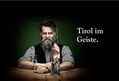 A man with a beard holds a long container from which a face appears. Next to him stands a bottle, and the text says "Tyrol in the Spirit".