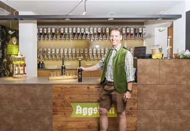 A friendly man in traditional Bavarian attire stands behind the bar. Behind him, numerous beer taps are lined up.