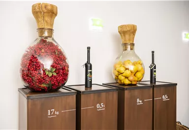 Three large glass carafes are on wooden pedestals. The carafes contain red and yellow fruits as well as bottles with liquids.