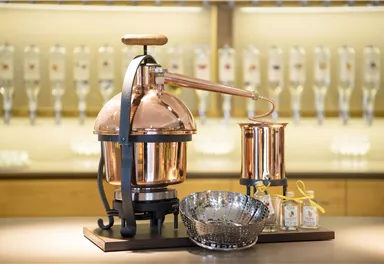 A copper distillation vessel stands on a table. In the background, several bottles are visible.