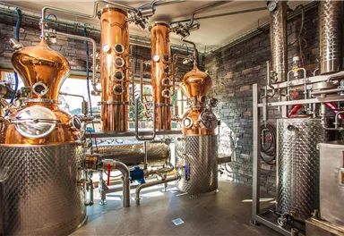 A modern distillery with shiny copper installations and stainless steel tanks. The walls are clad in stone and there is plenty of natural light.