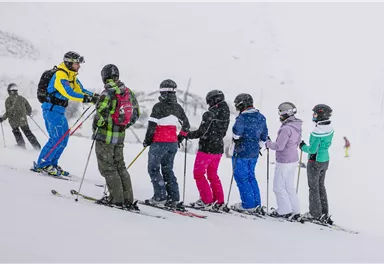 A group of skiers stands in a snowy landscape while receiving instructions from a teacher. It is snowing and the participants are wearing various ski clothing.